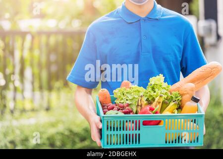 L'homme de livraison de magasin d'épicerie asiatique portant l'uniforme bleu et le masque facial protègent il livrant des légumes frais dans une boîte en plastique à la porte avant de la maison après Banque D'Images