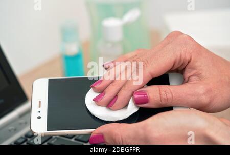 Femme nettoyant téléphone portable avec tampon en coton, essuyant écran noir, gros plan. Flacons flous et clavier d'ordinateur portable en arrière-plan. Banque D'Images