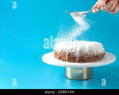 La main de femme saupoudrer le sucre glace sur un gâteau de petit pain frais fait maison. Le sucre en poudre tombe sur un gâteau au beurre frais et parfait sur fond bleu. Copier l'espace pour Banque D'Images