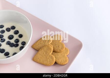 Petit déjeuner. Yaourt avec des biscuits en forme de cœur aux myrtilles. Ingrédients dans un plateau rose sur fond blanc. Détails du dessus avec espace de copie. Banque D'Images