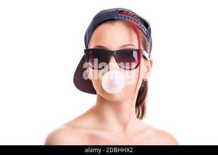 Belle fille élégante avec bouchon à dos et lunettes de soleil, soufflant une bulle rose avec sa gomme à mâcher, isolée sur fond blanc. Banque D'Images