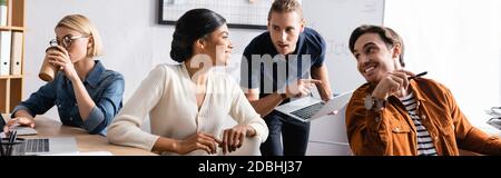 Souriante femme afro-américaine regardant des collègues tout en étant assise à un bureau près d'un collègue buvant du café dans le bureau, bannière Banque D'Images