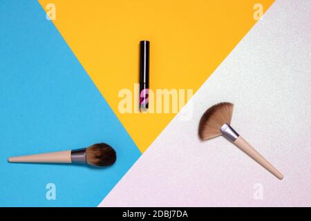 Deux pinceaux de maquillage professionnels et un rouge à lèvres plat reposent sur un fond rose vif, jaune et bleu avec espace de copie. Modèle pour les produits cosmétiques d Banque D'Images