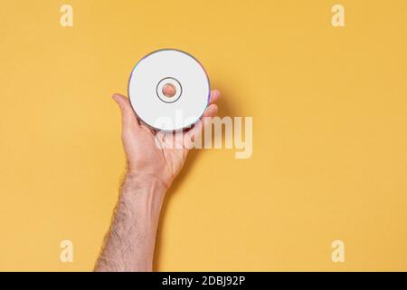 Main de sexe masculin tenant un CD-DVD devant un fond jaune, modèle de maquette modifiable pour votre conception, chemin de sélection d'étiquette inclus Banque D'Images