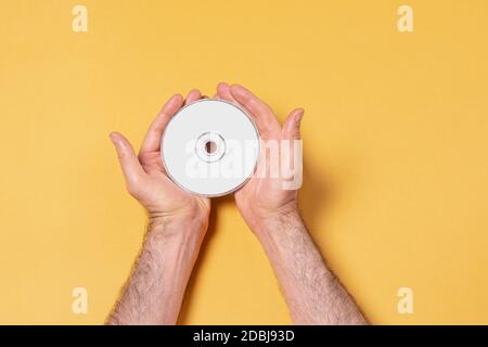 Main masculine tenant un CD-DVD par deux mains devant un fond jaune, modèle de maquette modifiable pour votre conception, chemin de sélection d'étiquette inclus Banque D'Images