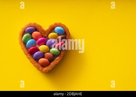 Boutons de friandises colorés sur gâteau en forme de coeur sur fond jaune - concept anniversaire Banque D'Images