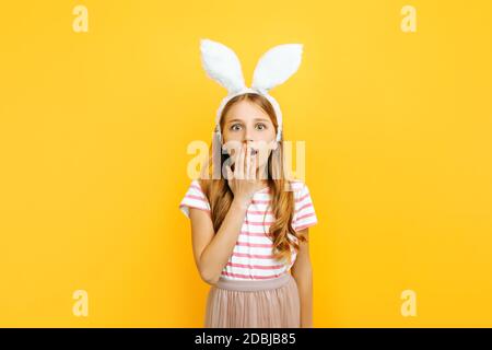 Choqué petite belle fille sur la tête avec des oreilles de lapin, surpris couvre sa bouche avec sa main sur un fond jaune. Symbole de Pâques et spri Banque D'Images