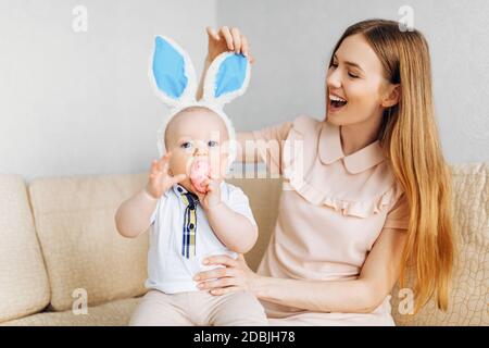Maman et bébé avec des oreilles de lapin, avec des œufs de Pâques dans leurs mains, assis sur le canapé, parents et enfants jouent à l'intérieur. La famille fête Pâques Banque D'Images
