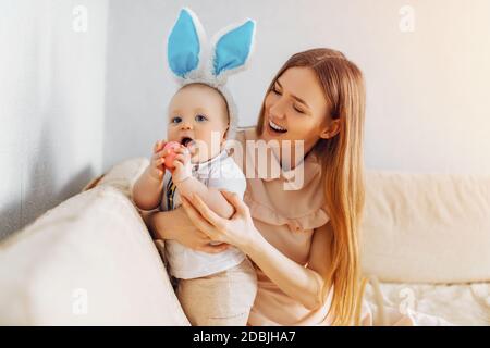Maman et bébé avec des oreilles de lapin, avec des œufs de Pâques dans leurs mains, assis sur le canapé, parents et enfants jouent à l'intérieur. La famille fête Pâques Banque D'Images