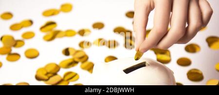 Mettez la pièce à la main à la tirelire. Banc de porc blanc sur fond de confetti de papier doré isolé sur fond blanc. Concept d'économie d'argent Banque D'Images