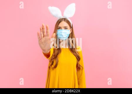 Une jeune fille dans les oreilles d'un lapin de Pâques porte un masque médical sur son visage contre les maladies virales et montre un geste d'arrêt, sur un bac rose isolé Banque D'Images