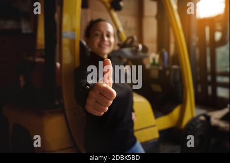 À l'intérieur de l'entrepôt, la jeune femme se tient devant un chariot élévateur jaune et montre ses pouces vers le haut. Accent sur le doigt, arrière-plan non mis au point. Banque D'Images