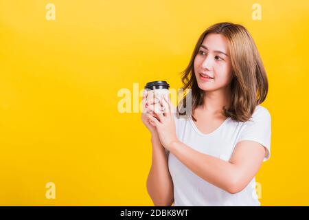 Portrait Asian Thai belle heureuse jeune femme porter un t-shirt blanc debout sourire tenant emporter café tasse en papier, studio tourné isolé sur le jaune Banque D'Images