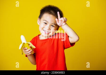 Joyeux portrait enfant ou enfant asiatique mignon petit garçon beau sourire portant un t-shirt rouge jouant tient pelé banane pour manger, studio tourné isolé sur Banque D'Images