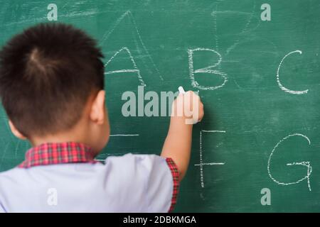 Rentrée des classes. Joyeux asiatique drôle de dos de mignon petit enfant garçon maternelle préscolaire dans l'uniforme d'étudiant écriture ABC avec craie blanche sur l'école verte Banque D'Images