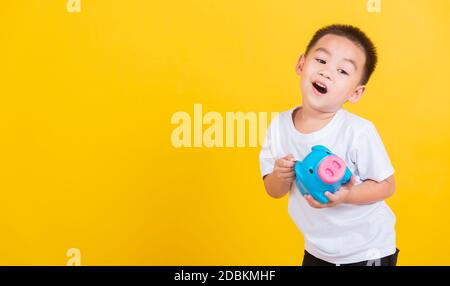 Asiatique Thai Happy portrait mignon petit gai jeune garçon sourire mettant de l'argent de pièce à la banque de piggy et regarder l'appareil photo, studio tourné isolé sur le jaune Banque D'Images