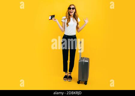 Une femme excitée et choquée, portant des lunettes de soleil avec une valise et un passeport, regarde la distance sur un fond jaune. Concept de voyage, Banque D'Images