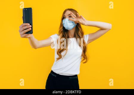 Une jeune femme dans un masque médical de protection prend un selfie sur un smartphone sur un fond jaune. Le concept de quarantaine, coronavirus Banque D'Images