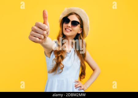 Joyeuse belle fille en lunettes de soleil et un chapeau d'été, montre un geste un pouce vers le haut, sur un fond jaune. Voyage, vacances, été Banque D'Images