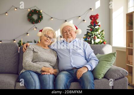 Joyeux couple senior en chapeau de père Noël regardant la caméra pendant assis sur un canapé à la maison Banque D'Images