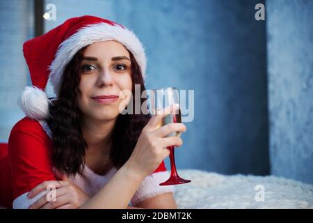 Jeune femme en costume du Père Noël avec une coupe de champagne couchée sur le lit. Charmante femme se reposant et célébrant Noël à la maison. Concept de vacances Banque D'Images