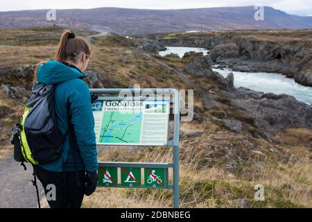 Une jeune femme portant une veste bleue vérifie le panneau Godafoss lorsqu'elle arrive à la célèbre cascade du nord de l'Islande. Banque D'Images