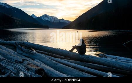 Un homme se tient sur une bûche et regarde le soleil se coucher sur le lac Duffey et le mont Matier, un jour d'automne en Colombie-Britannique. Banque D'Images