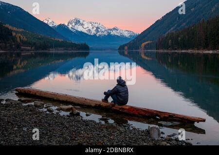 Un homme se tient sur une bûche et regarde le soleil se lever au-dessus du lac Duffey et du mont Matier le jour de l'automne en Colombie-Britannique. Banque D'Images