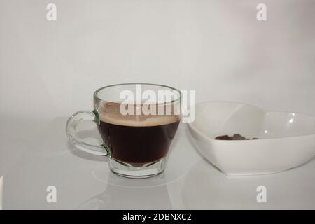Tasse à café en verre transparent contenant une boisson chaude caféinée prête à l'emploi avec mousse de café flottant à côté d'un bol en forme de cœur blanc contenant du rôti Banque D'Images