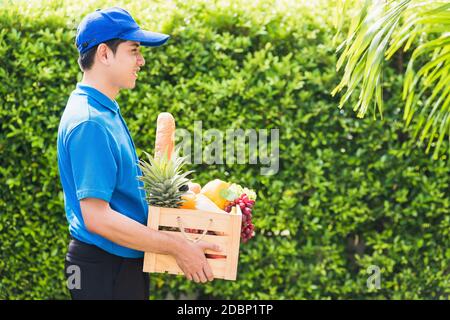 L'agriculteur asiatique porte l'uniforme de livraison il tenant les légumes et les fruits frais dans la caisse en bois dans les mains prêtes donner à la récolte biologique de client Banque D'Images