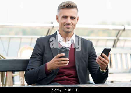 Le café me complète. Le boss tient le gobelet à emporter et le smartphone. Pause-café à l'extérieur. Boisson chaude. Technologie moderne. Petit-déjeuner le matin. Communication d'entreprise. Banque D'Images