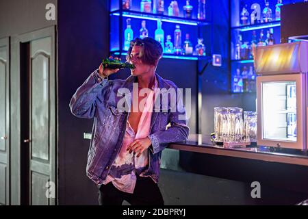 Un jeune homme boit une bière à l'intérieur d'un bar avec des néons en arrière-plan. Banque D'Images