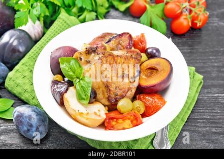 Poulet cuit avec tomates, pommes, prunes et raisins dans une assiette sur une serviette verte, ail, persil et basilic sur fond de bois foncé Banque D'Images