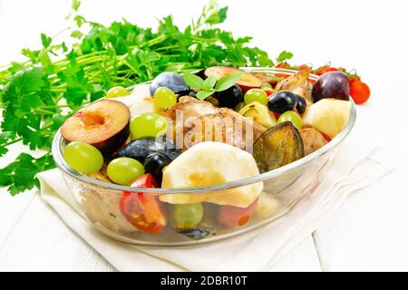 Poulet cuit avec tomates, pommes, prunes et raisins dans une rôtissoire en verre sur une serviette, ail, persil et basilic sur fond de bois clair Banque D'Images