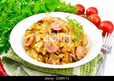 Ragoût de chou avec saucisses dans une assiette blanche sur une serviette, tomates, persil et fourchette sur un fond de bois Banque D'Images