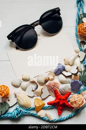 Carte blanche vierge avec espace pour le texte, lunettes de soleil, corde d'amarrage, coquillages et étoiles de mer rouges sur fond de tableau blanc en bois. Concept vacances d'été Banque D'Images