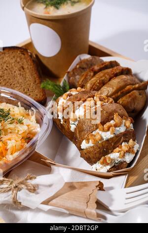 Vaisselle jetable, soupe dans une tasse de papier, pain, salade et pommes de terre cuites au four avec fromage cottage et noix sur un plateau en bois sur fond blanc. Blure Banque D'Images
