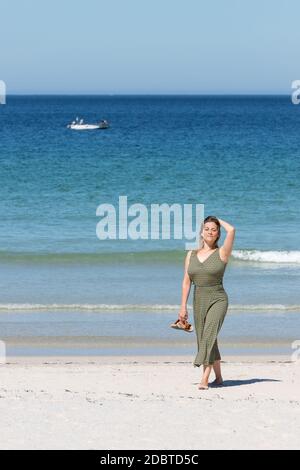 Femme blonde d'âge moyen vêtue d'une robe verte marchant pieds nus la plage sur la plage avec ses sandales en un main Banque D'Images