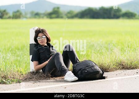 Young Asian woman cheveux courts et portant des lunettes de s'asseoir avec sac à dos randonnée le long d'une route de campagne à attendre de l'aide en Thaïlande Banque D'Images