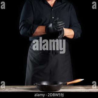chef en chemise et tablier noirs met des gants en latex noir sur ses mains avant de préparer les aliments, fond noir Banque D'Images