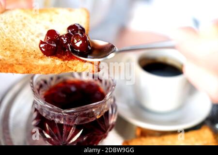 Petit déjeuner au lit dans l'hôtel. Main de femme qui répand de la confiture de cerise sur un toast. Banque D'Images