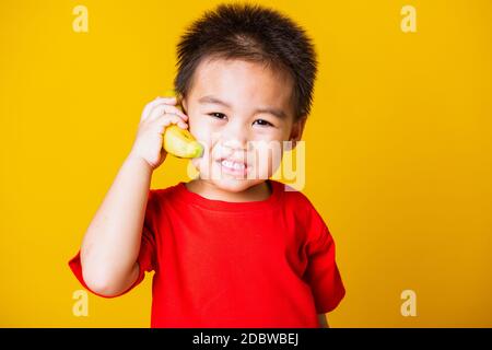 Joyeux portrait enfant ou enfant asiatique mignon petit garçon beau sourire portant un t-shirt rouge jouant tient banane fruit prétendant être comme un téléphone, s Banque D'Images