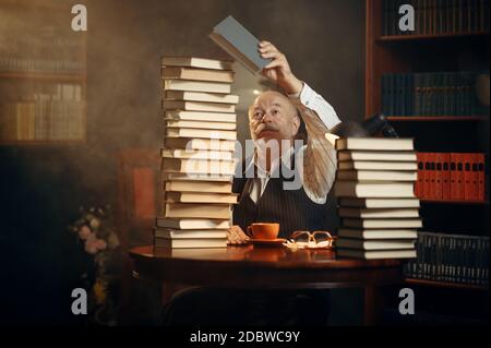 Un écrivain âgé lit à la table avec une pile de livres dans le bureau à domicile. Le vieil homme en verre écrit un roman de littérature dans la pièce avec de la fumée, de l'inspiration Banque D'Images