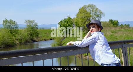 Portrait d'une femme russe aux cheveux rouges avec un maquillage brillant dans un chapeau et un chemisier bleu sur un fond de rivière dans la campagne. Copier l'espace Banque D'Images