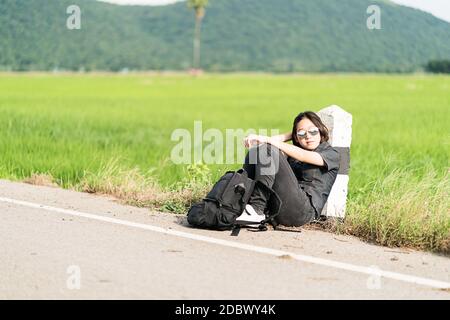 Young Asian woman cheveux courts et portant des lunettes de s'asseoir avec sac à dos randonnée le long d'une route de campagne à attendre de l'aide en Thaïlande Banque D'Images