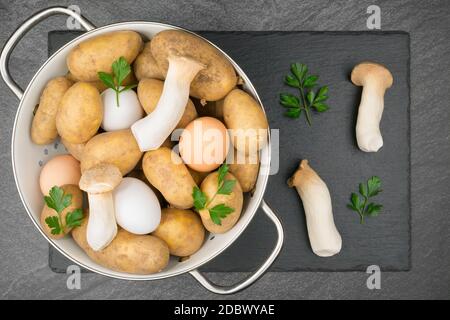 Pommes de terre crues, œufs, champignons des bois d'huîtres et feuilles de persil dans un tamis en émail blanc sur une laque d'ardoise. Pose à plat. Banque D'Images