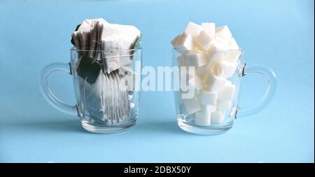 Deux grandes tasses en verre transparent pleines de sucre blanc raffiné et sachets de thé sur fond bleu. Concept de thé à boire fréquemment avec du sucre comme danger pour la santé Banque D'Images