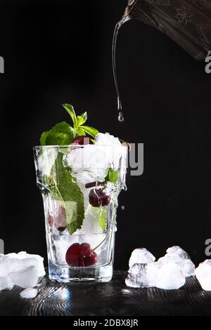 La limonade fraîche est versée à partir d'un vaisseau d'époque dans un verre avec des cerises et de la glace. Feuilles de menthe dans un verre. Boisson d'été en saison. Photo sur fond noir Banque D'Images