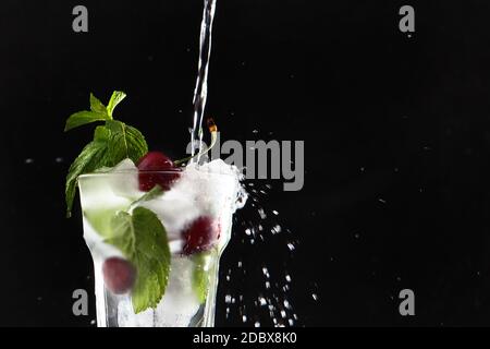 La limonade fraîche est versée dans un verre avec des cerises et de la glace. Feuilles de menthe dans un verre. Boisson d'été en saison. Photo sur fond noir. Copier l'espace. Banque D'Images