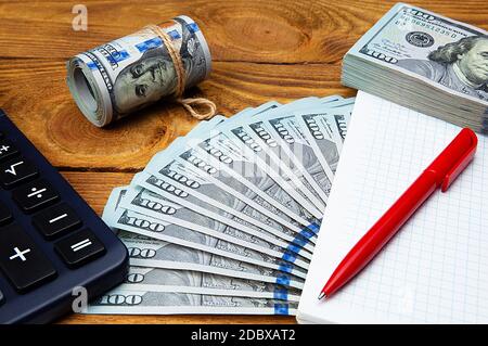 Gros plan de dollars américains répartis et en rouleau, un carnet d'affaires vide, un stylo et une calculatrice sur une table en bois de couleur claire. Banque D'Images